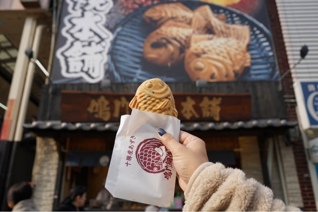 Woman's hand holds up a taiyaki in front of a taiyaki shop in Osaka.