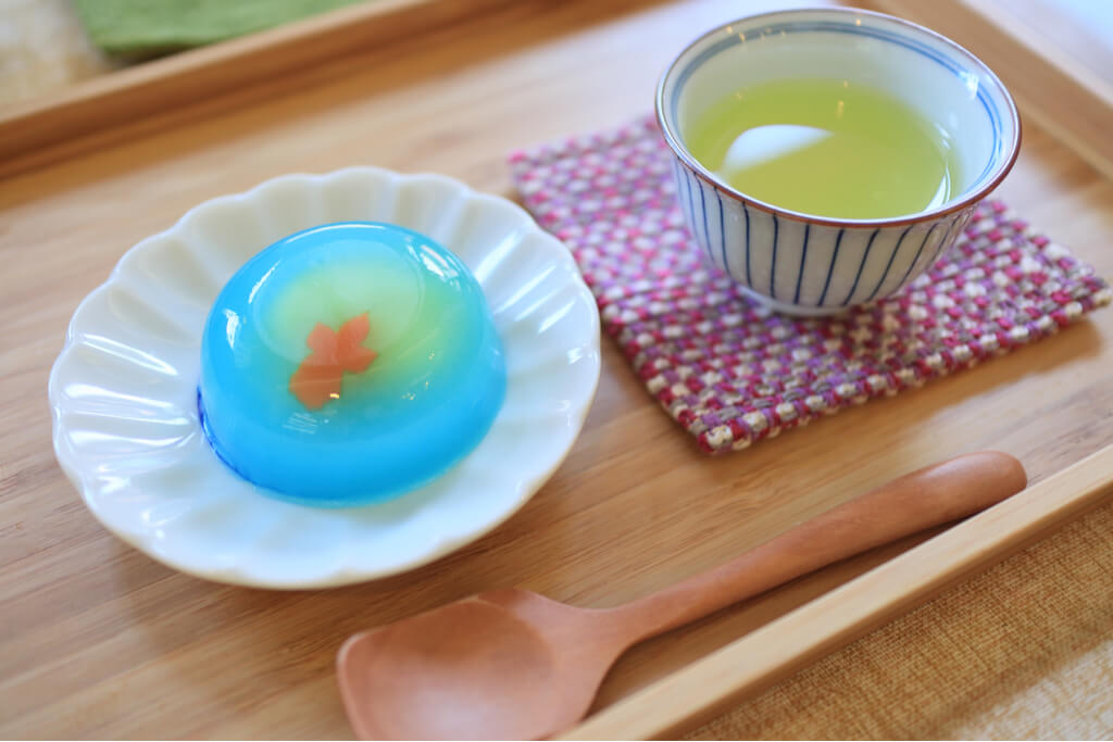 Mizu yokan with a mini goldfish made of read bean paste next to a cup of cool green tea and a wooden spoon