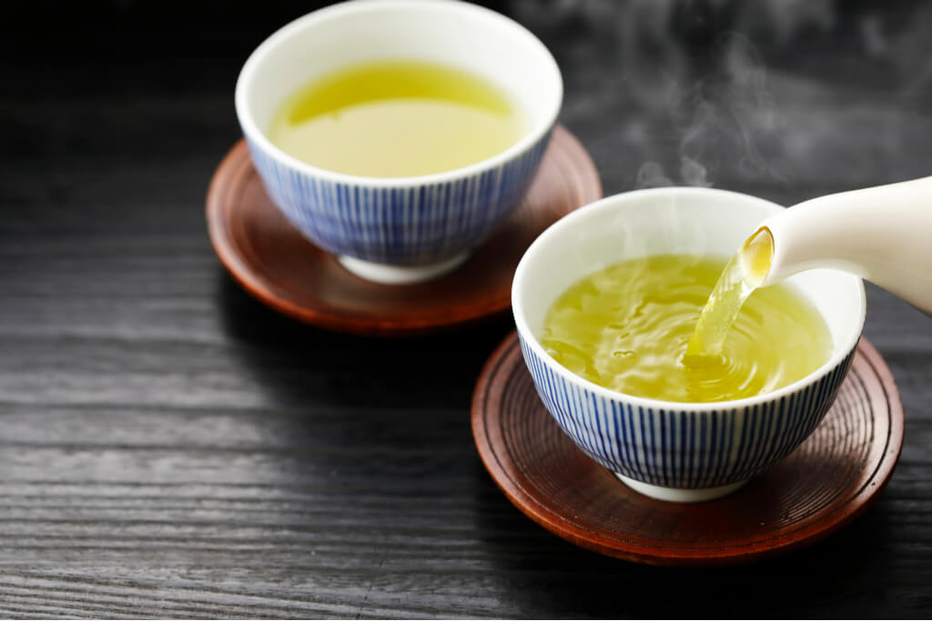 Two cups of sencha, one being poured from a white pot. There are many sencha tea benefits.