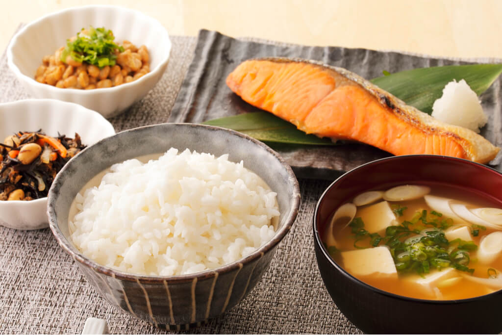 A Japanese breakfast of a bowl of rice, miso soup, salmon, natto, and vegetable on a gray placemat.