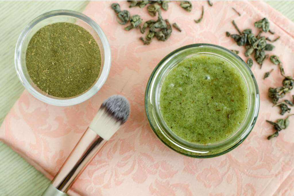 A bowl of powdered green tea, AKA matcha, next to a larger bowl of matcha exfoliant on a pink cloth on a green table with a makeup brush.