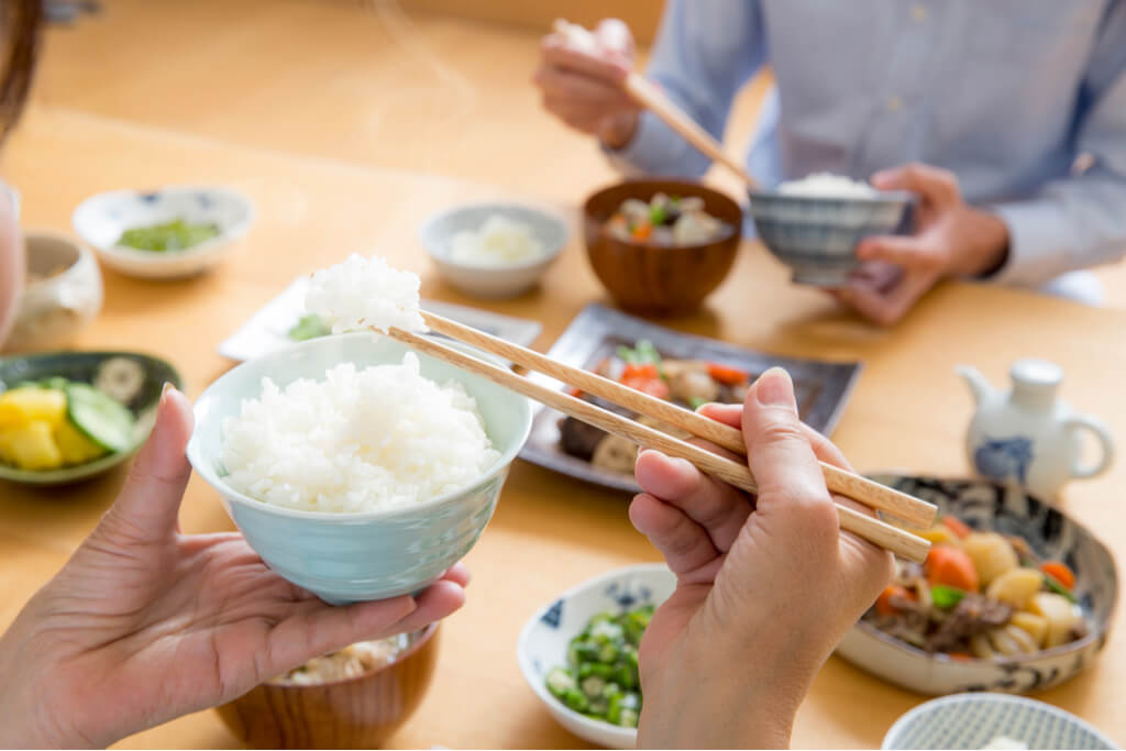 A woman hands holding rice in front of a spread of Japanese food with another person sitting across also holding rice.