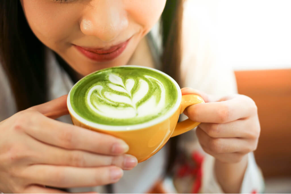 A woman holds and smells a matcha latte with latte art in a yellow mug.
