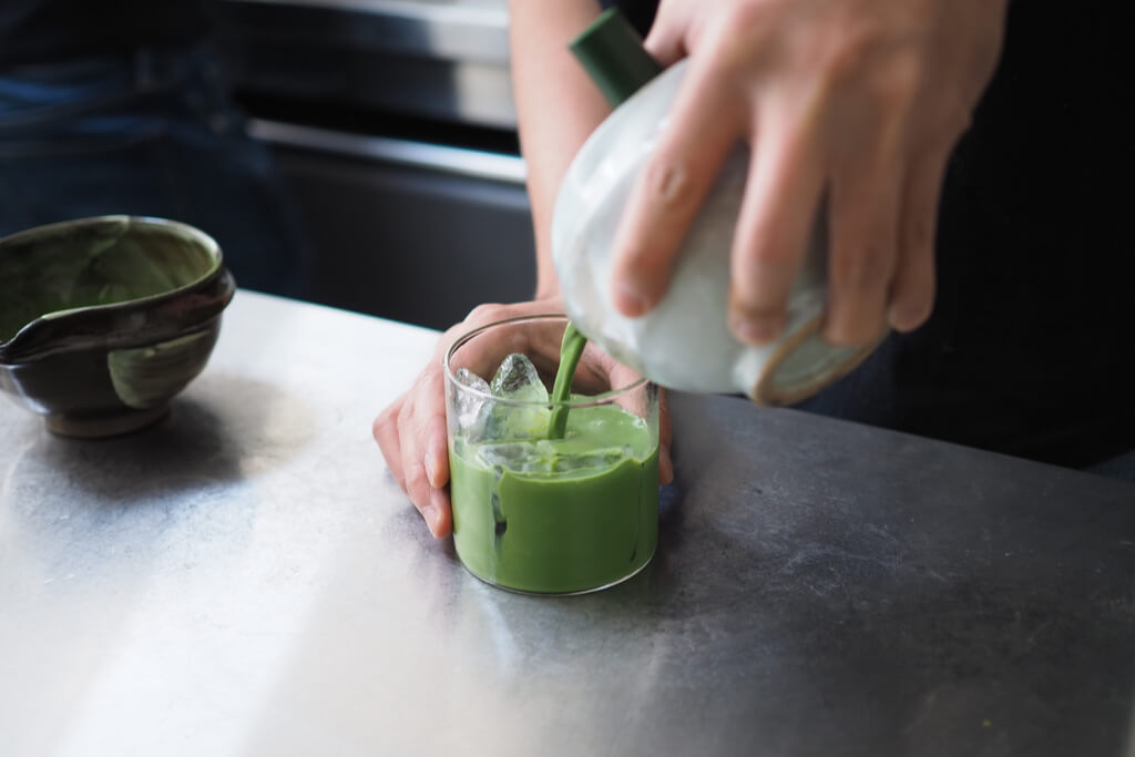 Hands pour matcha from a traditional matcha bowl into a glass of ice.