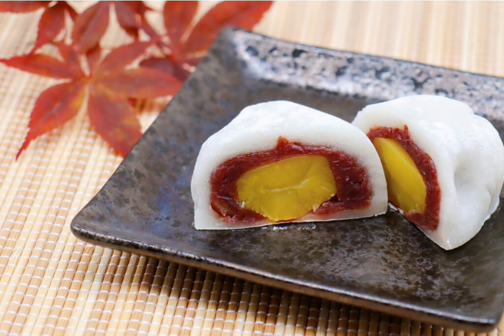 A manju made with a mochi outer, stuffed with chestnut and red bean paste on a Japanese ceramic plate on a bamboo mat with red leaves behind it.