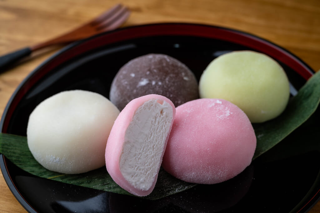 Five pieces of mochi ice sit on top of a leaf on a circular Japanese plate on a table with a wooden dessert fork in the background.