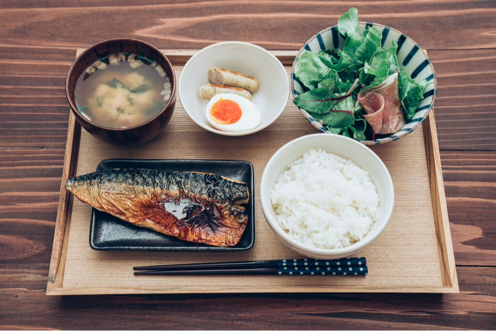 A Japanese breakfast of grilled mackerel, rice, miso soup, a salad with ham, half a boiled egg, and sausage on a wooden tray on a wood table.