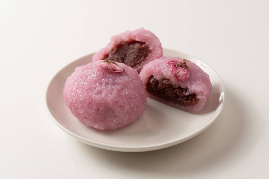 Two sakura manju sit on a white plate on a white table, with one split open revealing the red bean paste center.