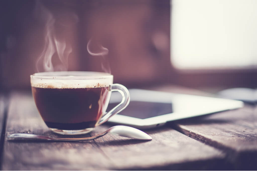 A steaming hot cup of coffee sits on a wooden table with a spoon in front of it and an ipad next to it.