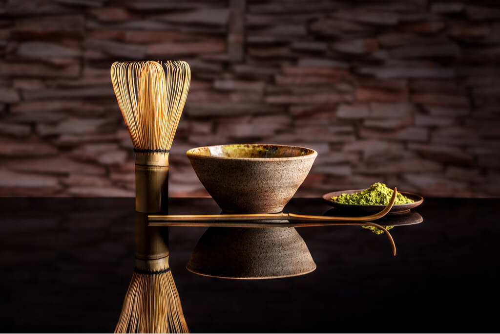 A raku ware bowl in between a green tea whisk, a plate of green tea, and a green tea powder scooper.