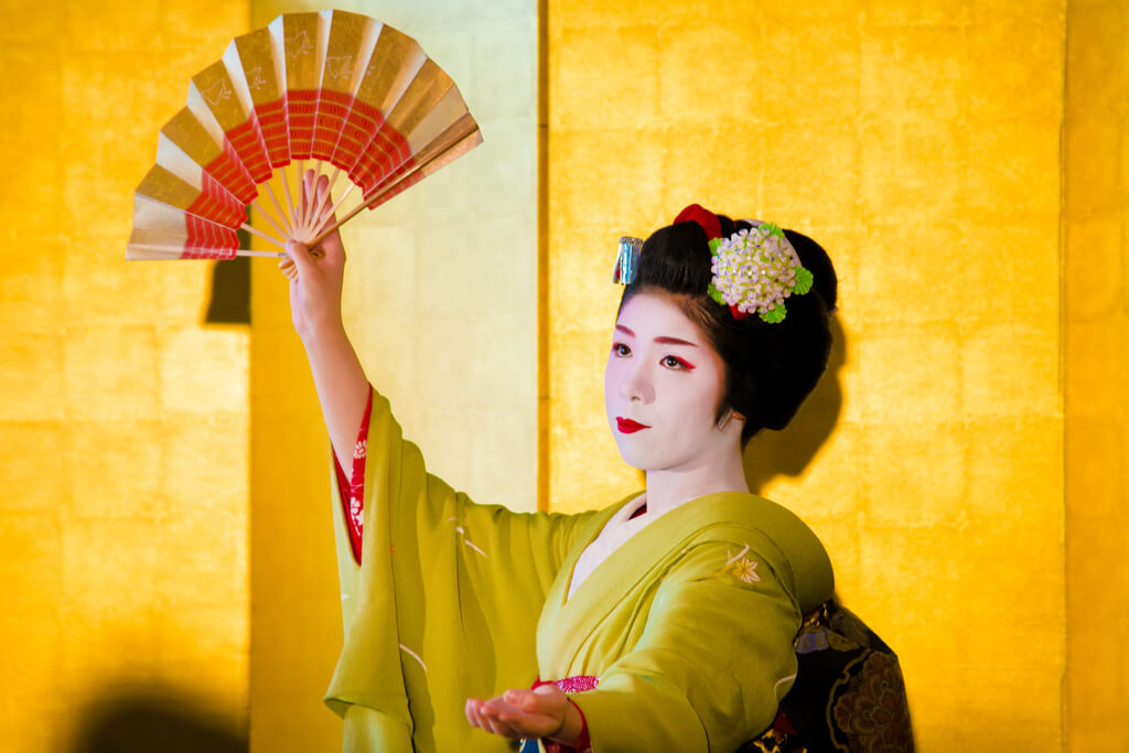A geisha in a green kimono does a traditional dance with a gold and red fan in front of a gold background.