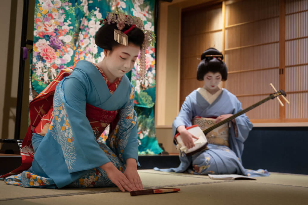 Two geisha women sit on a bamboo mat floor during a performance where one is playing the shamisen and the other is bowing in front of a fan.