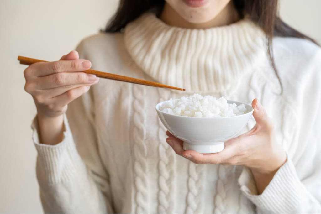 A woman in a white sweater holds a small bowl of rice in one hand and chopsticks in the other