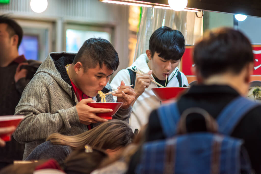 Two men stand in a busy ramen shop and eat noodles out of a red bowl.