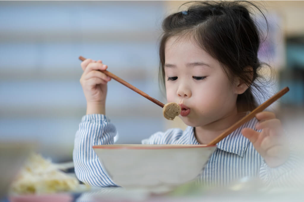 A little girl blows onto some food that she has stabbed a chopstick through over a bowl of the food.