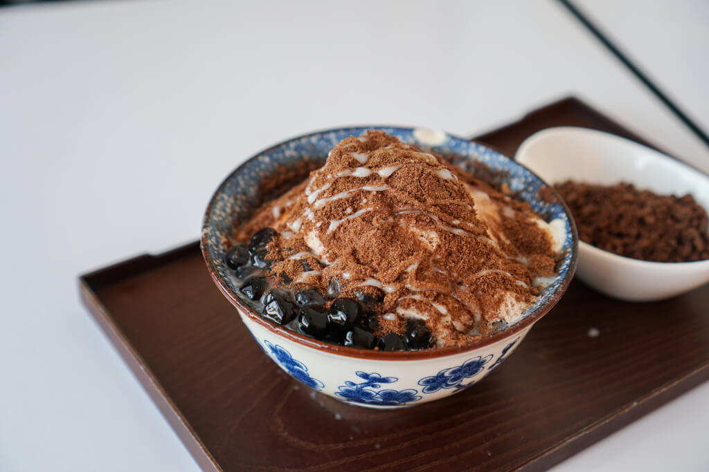 Tofu pudding with condensed milk, cocoa powder, and tapioca balls in a white and blue bowl on a tray next to a bowl of cocoa powder on a white background