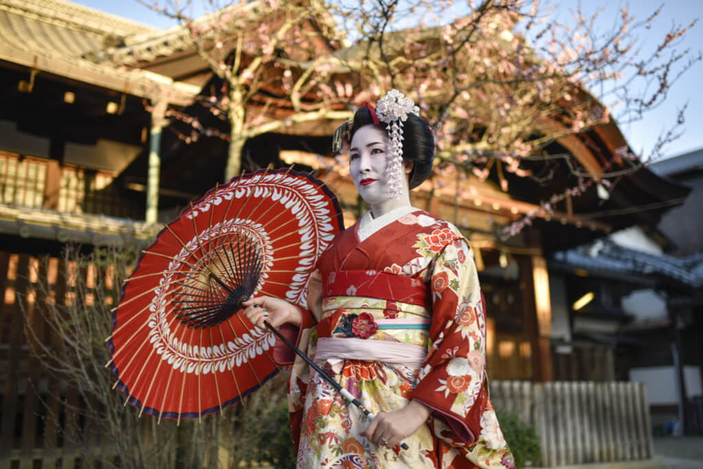 A full-fledged geisha walks down the street in a red kimono with a red parasol in front of a traditional Japanese building.