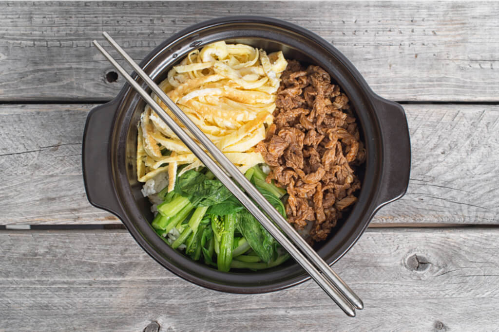 A bowl of Korean food with beef and vegetables with a pair of Korean metal chopsticks laid over it.
