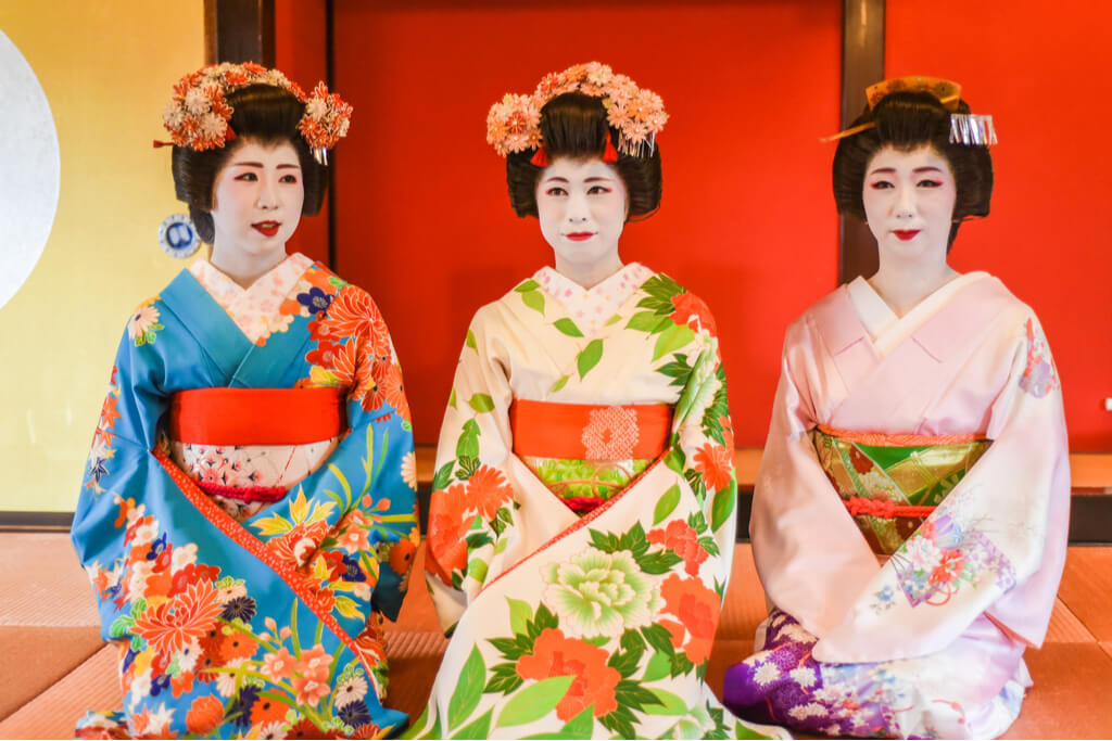 Three different maiko sit on the floor in front of a red door on tatami, with colorful kimonos.