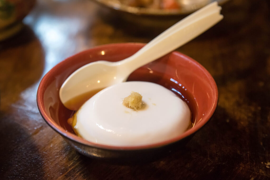 A bowl of tofu pudding covered in a sauce with a plastic spoon stuck into it on a dark table with food in the background