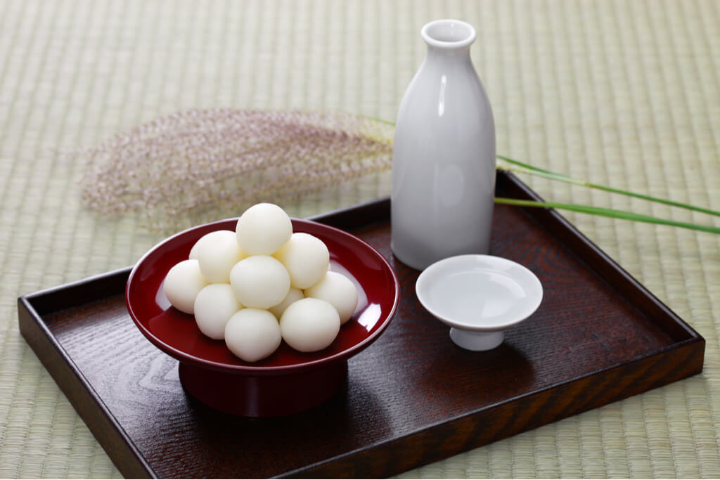 A bowl of tsukimi dango in a red container on a wooden tray that also holds a white sake bottle, white sake cup, and a piece of rice plant on a tatami floor.