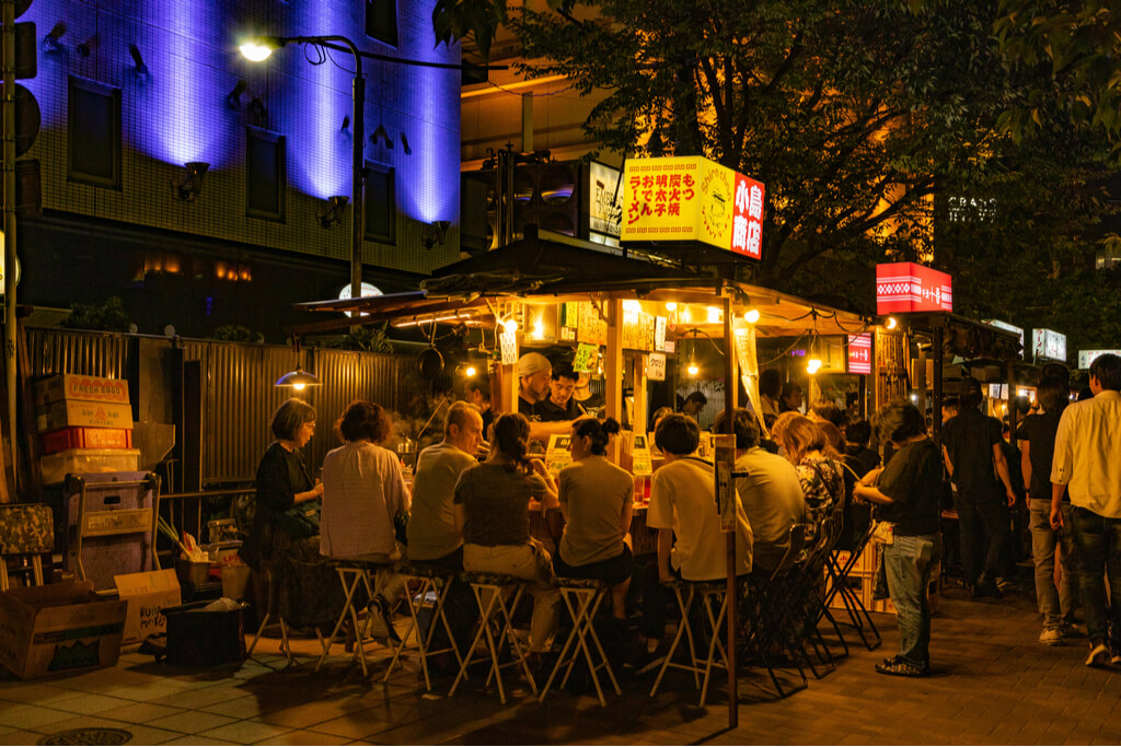 A food stand or yatai in Fukuoka serving Kyushu ramen to customers sitting around the stand on a busy street.