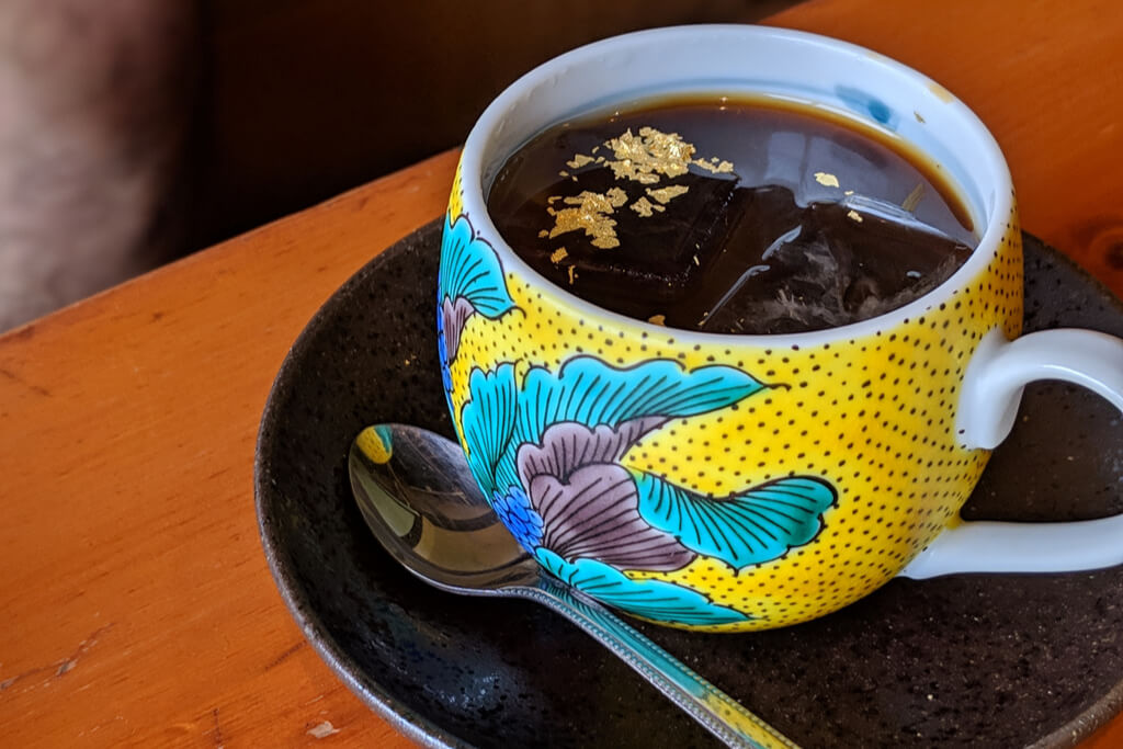 A colorful yellow, blue, green, and purple kutani cup filled with coffee with gold leaf sprinkles on a black plate on a wood table.