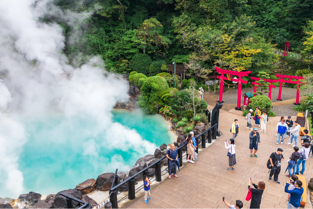 Many people taking pictures in front of a blue pool of hot spring water with steam coming from it and some torii gate and a lot of greenery behind it.