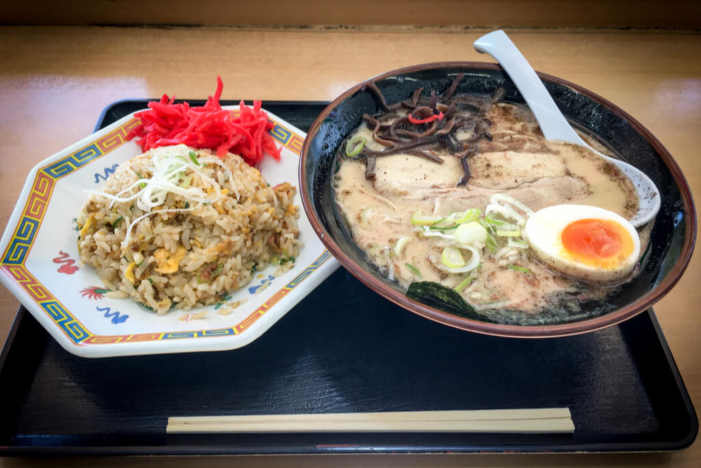 A bowl of Hakata style Kyushu Ramen with black pepper in the broth next to a plate of fried rice with red pickled ginger on a black tray on a counter