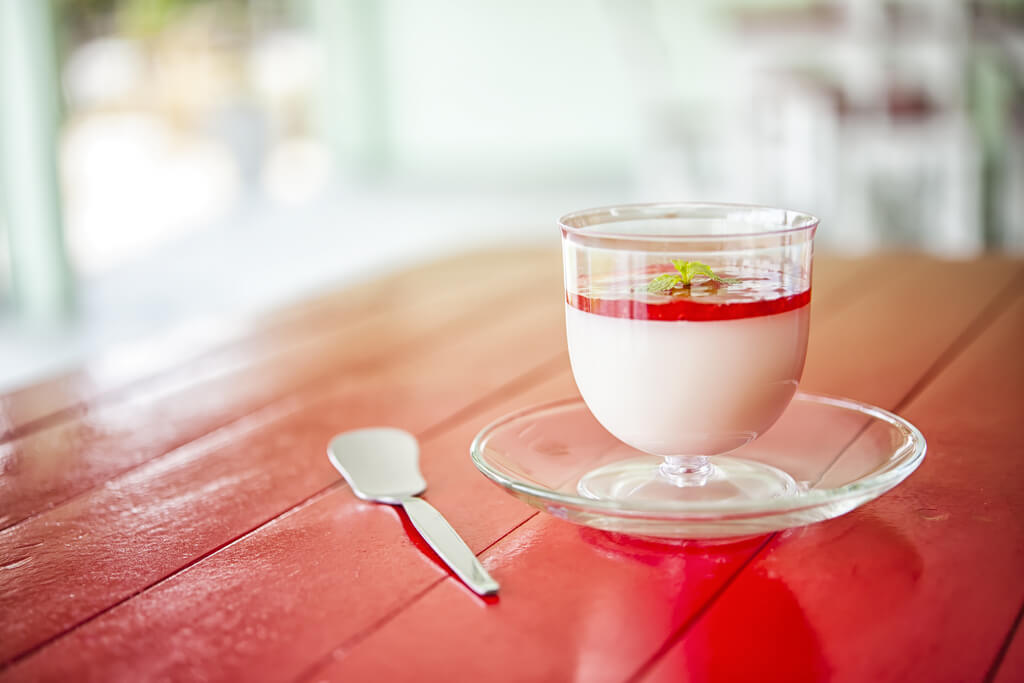 A cup of milk pudding with a fruit jelly topping on a plate next to a spoon on a red table