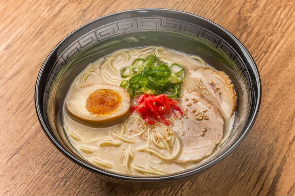 A bowl of Hakata ramen with half of a seasoned egg, green onions, pickled ginger, and pork belly in a black bowl on a wooden table