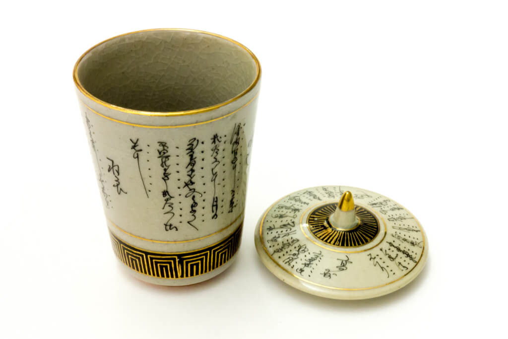 A beige kutani cup featuring traditional Japanese writing with black and gold accents on a white background 