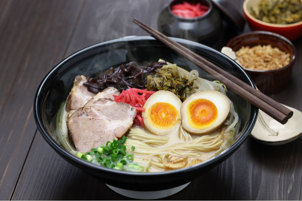 A bowl of Kyushu ramen with egg, pickled ginger, pork, and other toppings with extra toppings in the background.