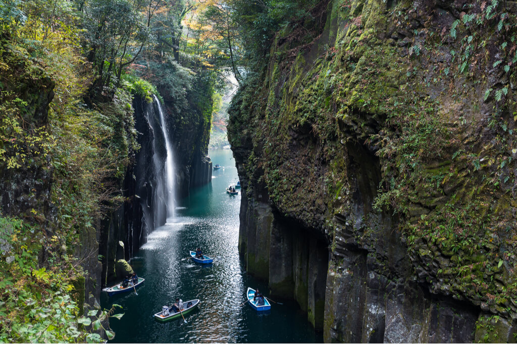 People in rowboats on the river sandwiched between the steep sides of the Takachiho Gorge with a waterfall on the left side of the gorge.