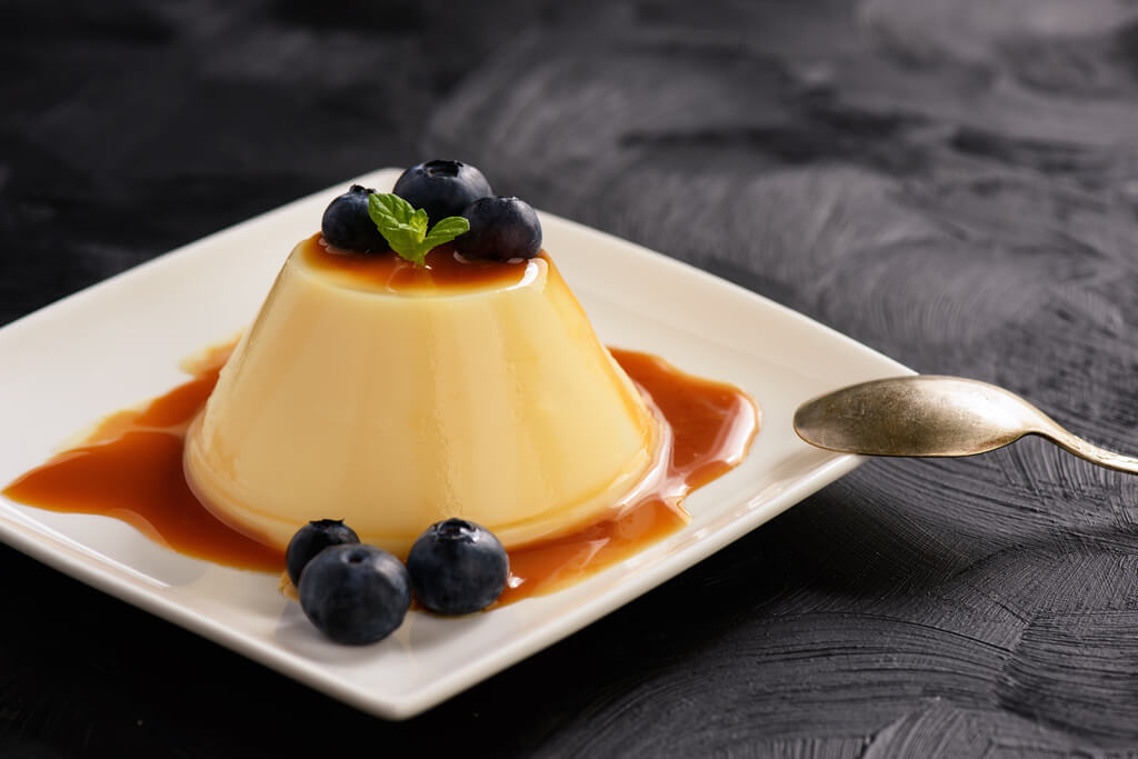 Japanese custard pudding topped in a caramel sauce with blueberries and a little mint on top on a white plate on a black background