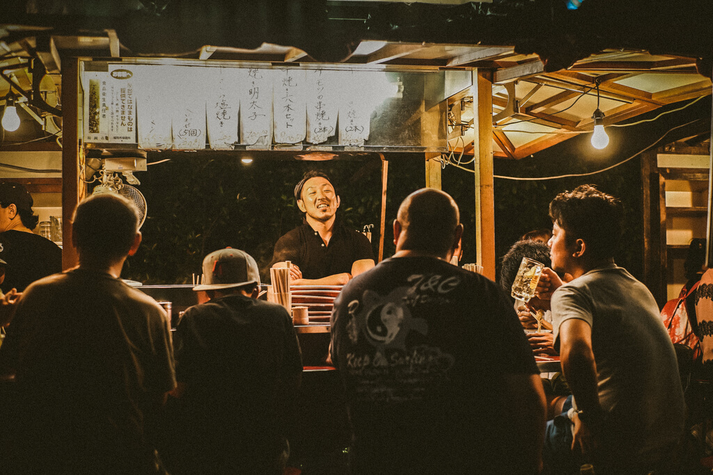 A food stall with the owner standing behind it while talking with the customers sitting at the bar drinking and eating ramen