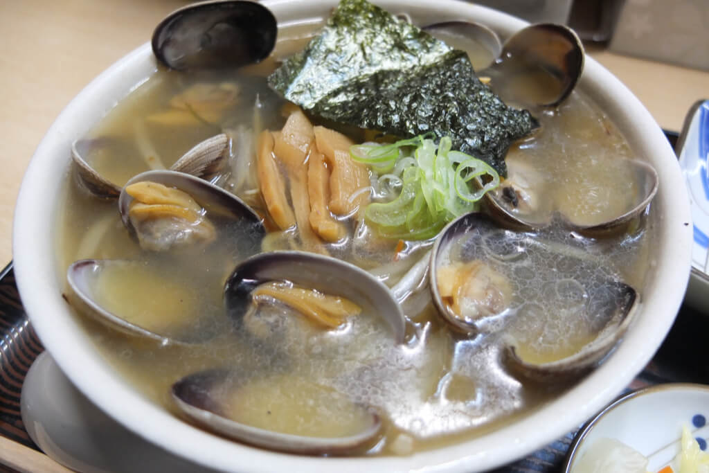 A bowl of Kushiro ramen, a Hokkaido ramen type, with a clear broth with clams, bamboo, onion, and seaweed over the noodles.