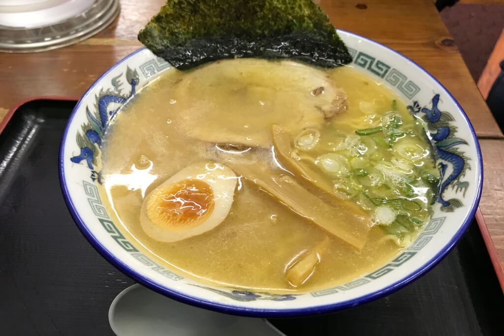 A bowl of Asahikawa ramen, a Hokkaido ramen type, topped with an egg, grean onions, bamboo shoots, chashu, and seaweed, on a tray in a restaurant.