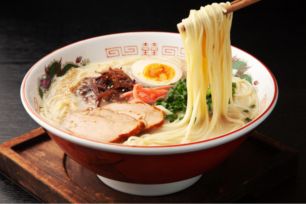 A pair of chopsticks picking up noodles from a bowl of Hakata ramen, a famous Kyushu local food.
