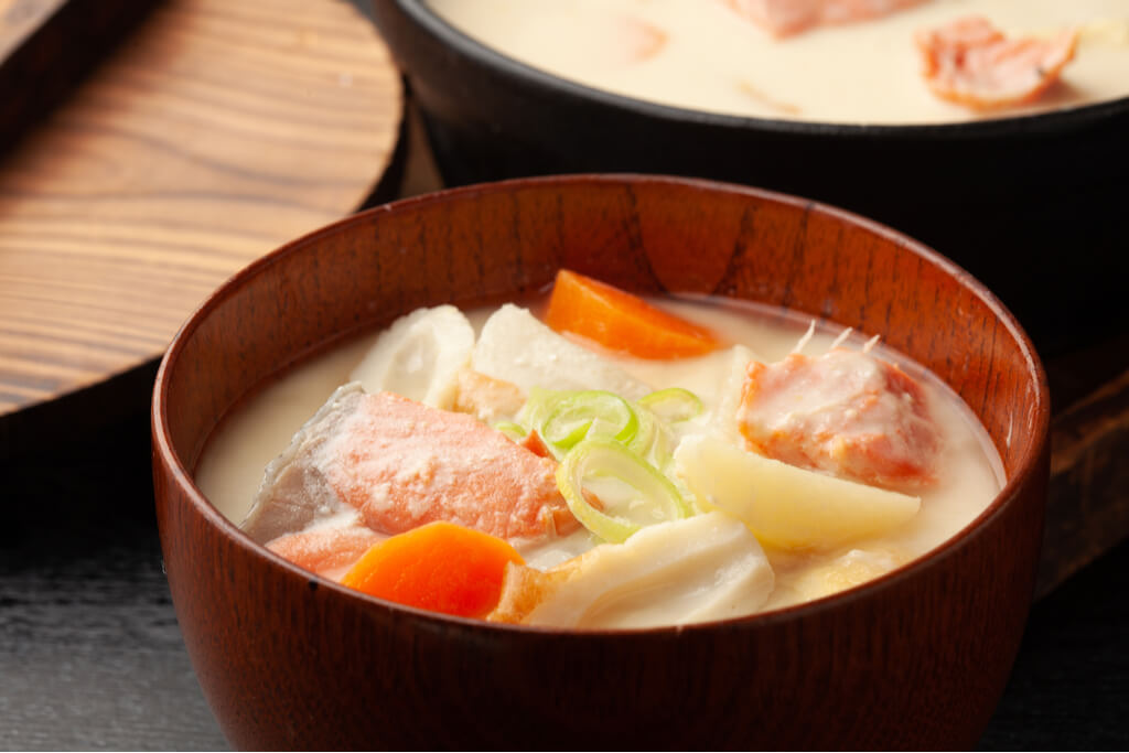 A wooden bowl full of Ishikari nabe made with salmon, carrots, and more, in front of the larger pot