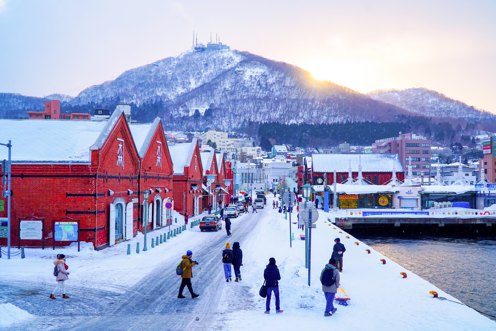 the sun rises over a mountain in Hokkaido. There are people outside next to red houses and snow is everywhere