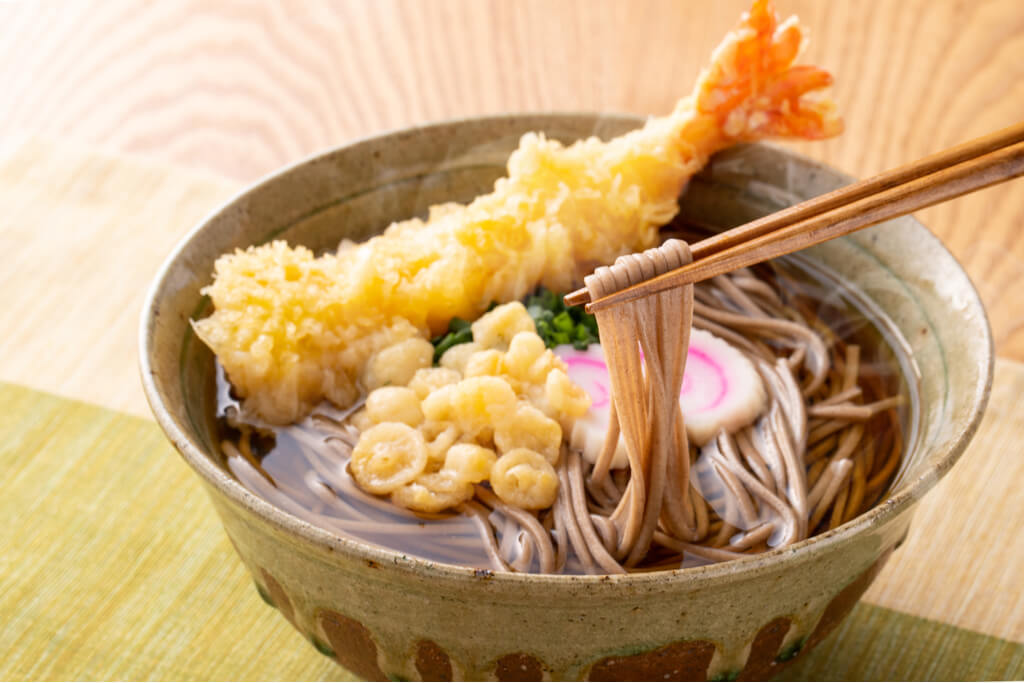 A pair of chopsticks pulls noodles out of a bowl of toshi-koshi soba, a Japanese new year food, with tempura shrimp on top.