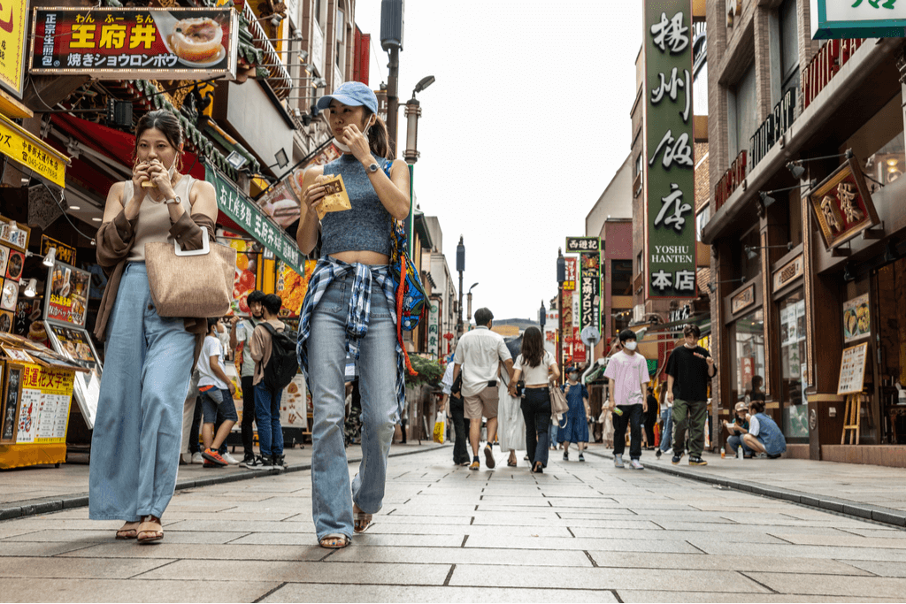 Two women walk down the street of the Yokohama Chinatown while eating some food from a street vendor with many people in the background.