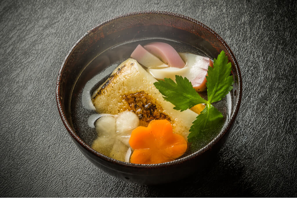 A bowl of ozoni, a Japanese new year soup, with grilled mochi, carrots, and other vegetables on a black background.