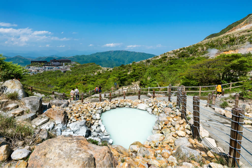 People walk on a mountain hiking trail with a sulfur hot spring in the foreground and a popular Hakone onsen, Kowakudani, in the background.