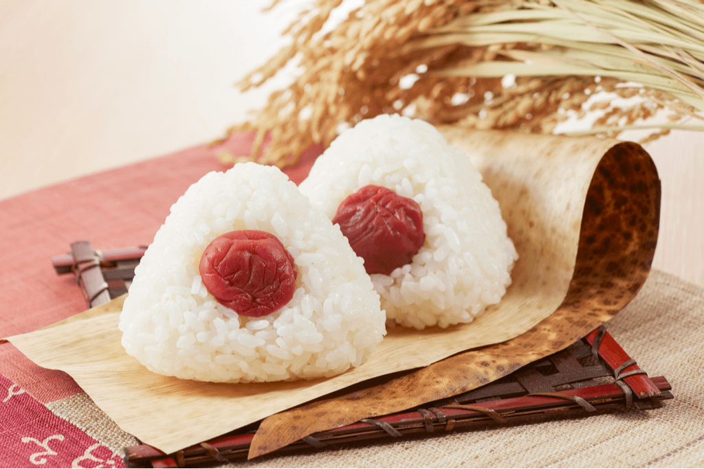 Two Japanese rice balls on a paper and serving plate with Japanese umeboshi on them and dried rice plants in the background