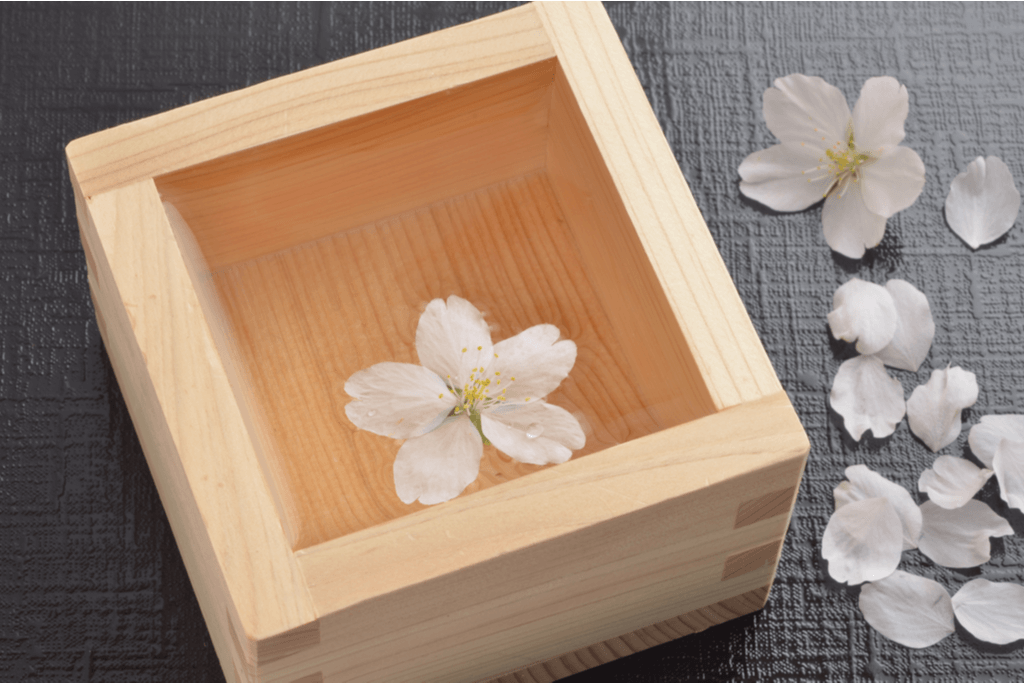 A wooden box used as a cup for Japanese sake sits on a black table with a cherry blossom sitting on top of the drink.