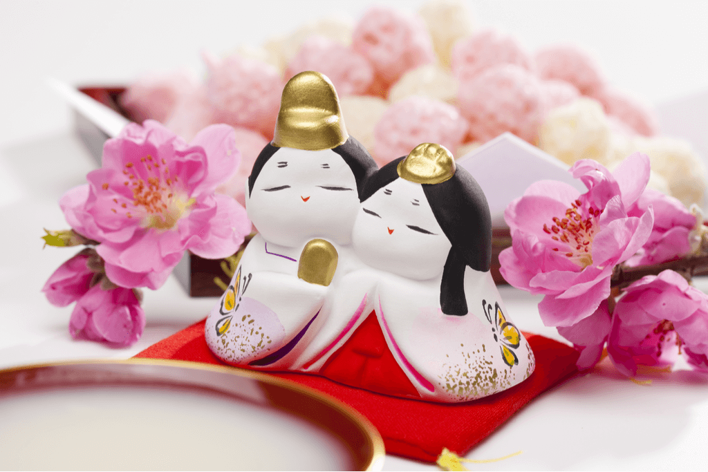 A figure showing two Hina characters with a bowl of colorful, puffy arare on a white plate behind it with plum blossoms on either side of the figures.