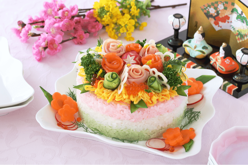 A chira sushi, a type of sushi served on Hinamatsuri, sits on a white plate with rice in layers of green, pink, and white, and lots of fish and vegetables arranged on top.