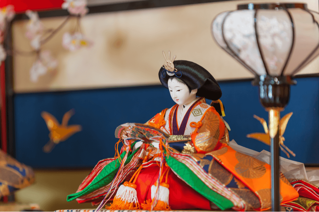 The princess doll, one of the most important of a Hina dolls set, sits with a fan in a colorful royal kimono in front of a screen and behind a lamp with a golden crane.